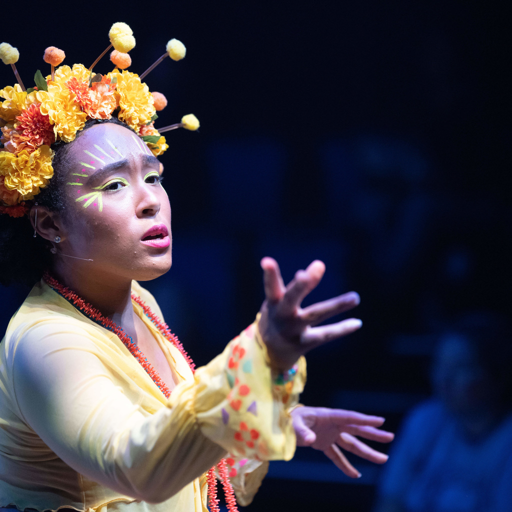 A young woman wearing a yellow dress and a crown of yellow flowers gestures toward an audience.