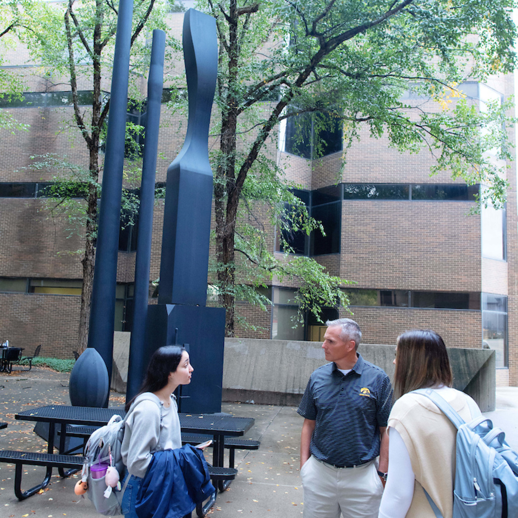 three people standing in the courtyard of a building with a green tree and a black sculpture in the background