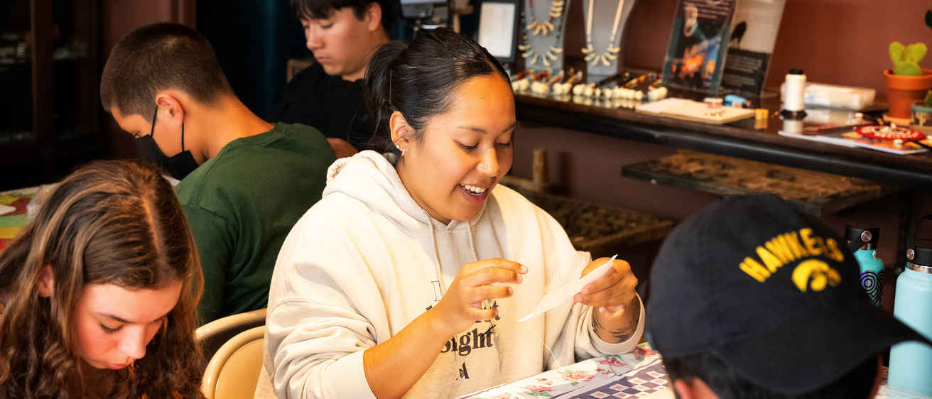 A group of teenagers works on projects around a table. A smiling girl in a white hooded sweatshirt is at the center of the photograph.