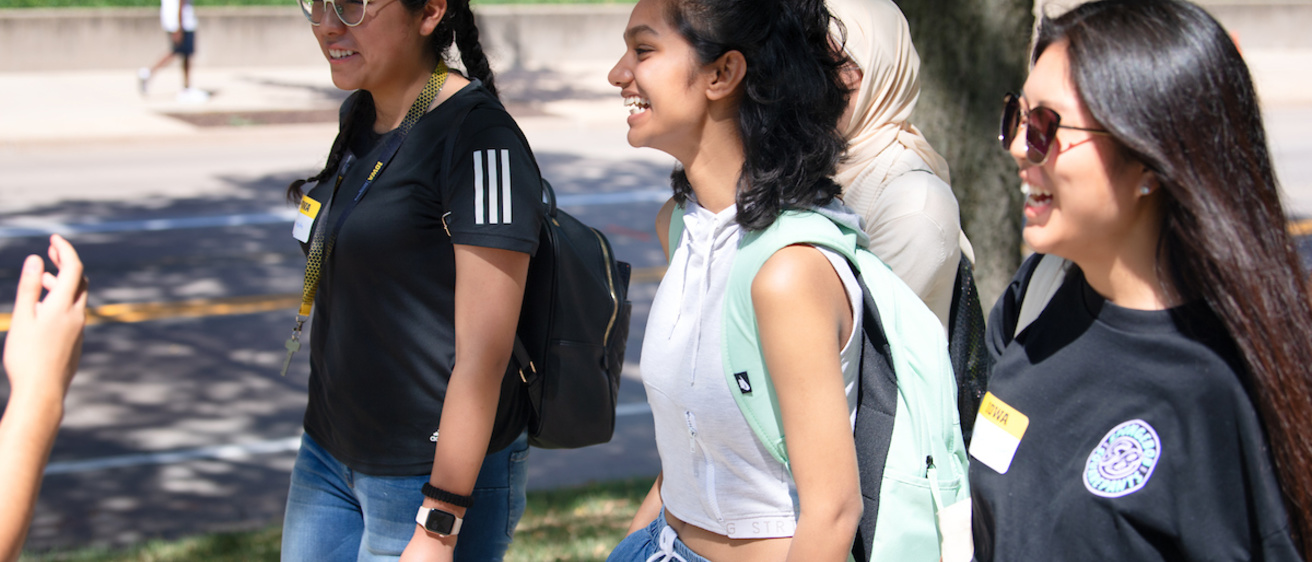 Three young women dressed in warm weather clothing and sunglasses walk down a street.