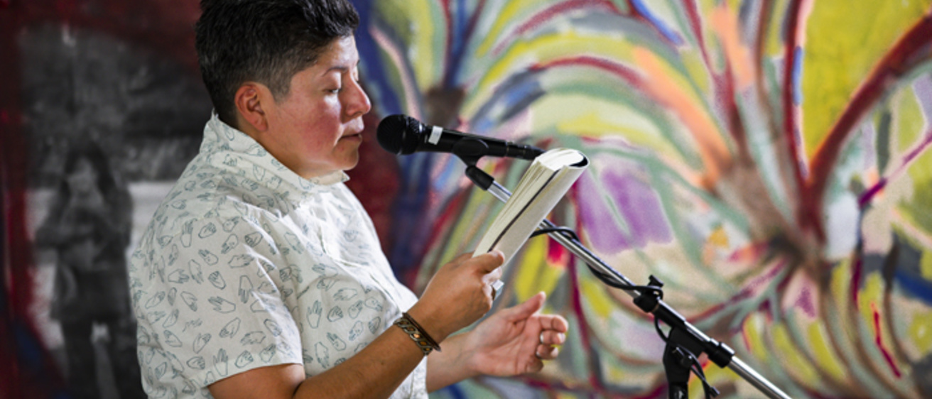 Middle aged woman with dark brown hair in a shirt short sleeved shirt is holding a book and standing at a microphone reading. There is a colorful mural in the background.