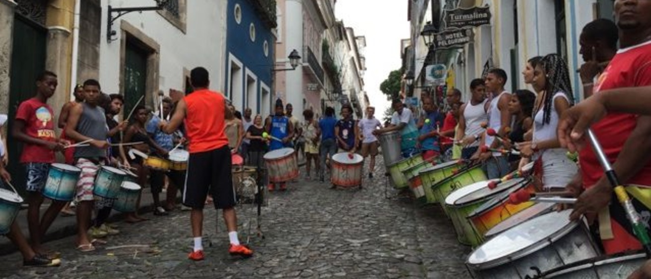 A group of men with drums around their necks stand in a cobblestone street.